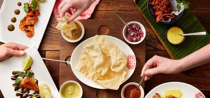Papadums are being grabbed and dipped into sauces and chutneys, while surrounding plates of Indian appetizers sit on a wooden table, creating a shared meal setting.