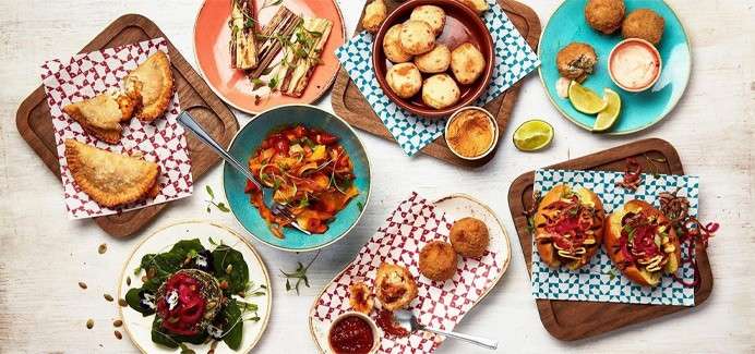 Assorted tapas sit arranged for sharing, including empanadas, arancini, grilled vegetables, salad, soup, and sliders, with dipping sauces and lime wedges on colorful plates and patterned papers over wooden boards.