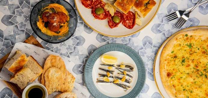 Dishes sit arranged on a patterned table: pizza, anchovies with lemon, salami with focaccia and olives, fried croquettes on sauce, and breads with olive oil; paper liner shows illegible brand text.