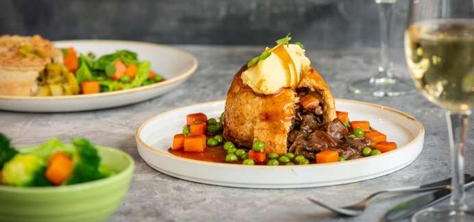 Meat-filled pastry sits cut open, releasing stew, topped with mashed potato and gravy; peas and diced carrots ring it. On a tabletop, wine glass, side salad, and broccoli bowl surround.