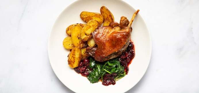 Roasted poultry leg rests on a white plate, accompanied by golden roasted fingerling potatoes, sautéed spinach, and a dark fruit sauce, presented on a light marble surface.