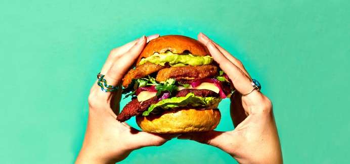 Burger is held by two hands; layers include fried cutlets, lettuce, red onion, cheese, and guacamole; set against a solid teal background.
