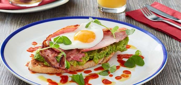 Open-faced toast topped with mashed avocado, bacon, and sunny-side egg rests on white plate, drizzled with red sauce, garnished with greens; set on wooden table with juice, cutlery, napkin.