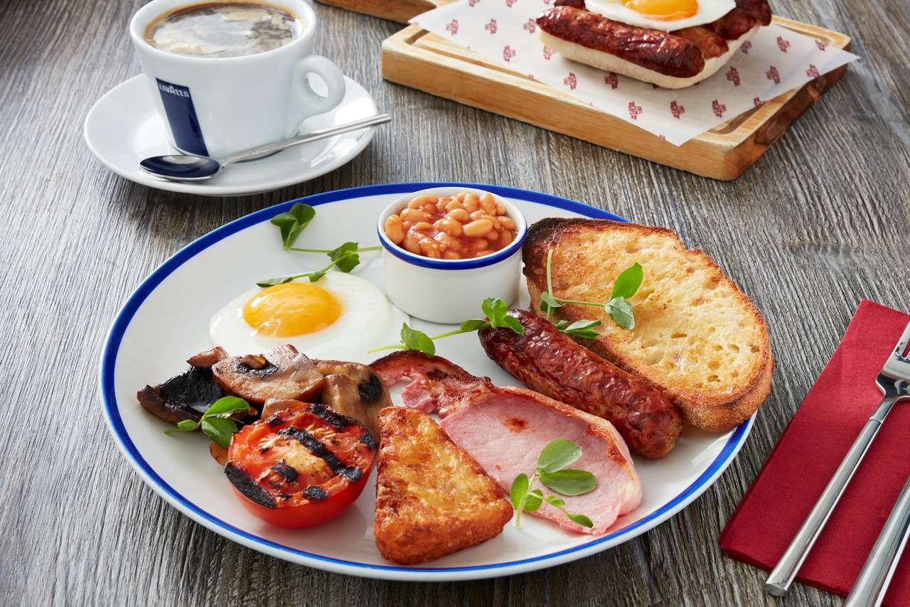 Open-faced toast topped with mashed avocado, bacon, and sunny-side egg rests on white plate, drizzled with red sauce, garnished with greens; set on wooden table with juice, cutlery, napkin.