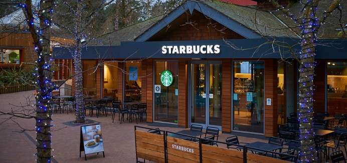 Starbucks café illuminates its wooden storefront; interior counter glows through glass doors. Empty patio tables surround it under blue-lit trees in a plaza. Text: STARBUCKS; STARBUCKS.