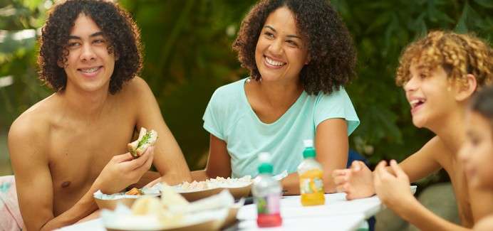 Group of four people sit around a table, smiling and chatting while eating snacks and sandwiches; drinks and bowls of food on the table; leafy garden background.