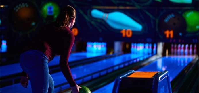 Bowler grips a green ball and prepares to roll; neon-lit lanes lead to standing pins, with large glowing lane numbers “10” and “11” and bowling graphics illuminating the dark alley.