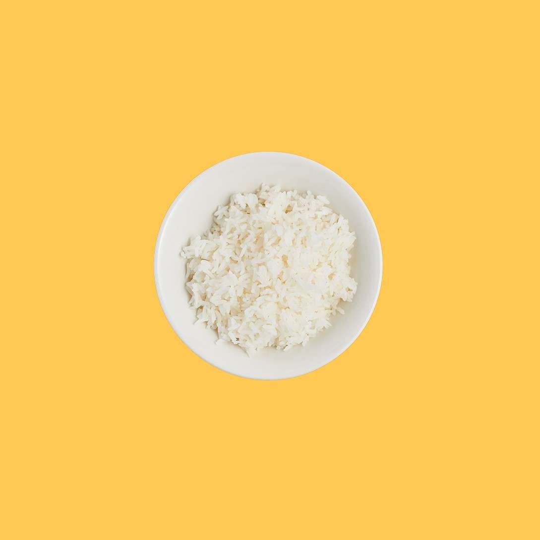 White bowl of fluffy cooked rice sits centered, resting still against a solid mustard-yellow background, creating a clean, minimalist food presentation.