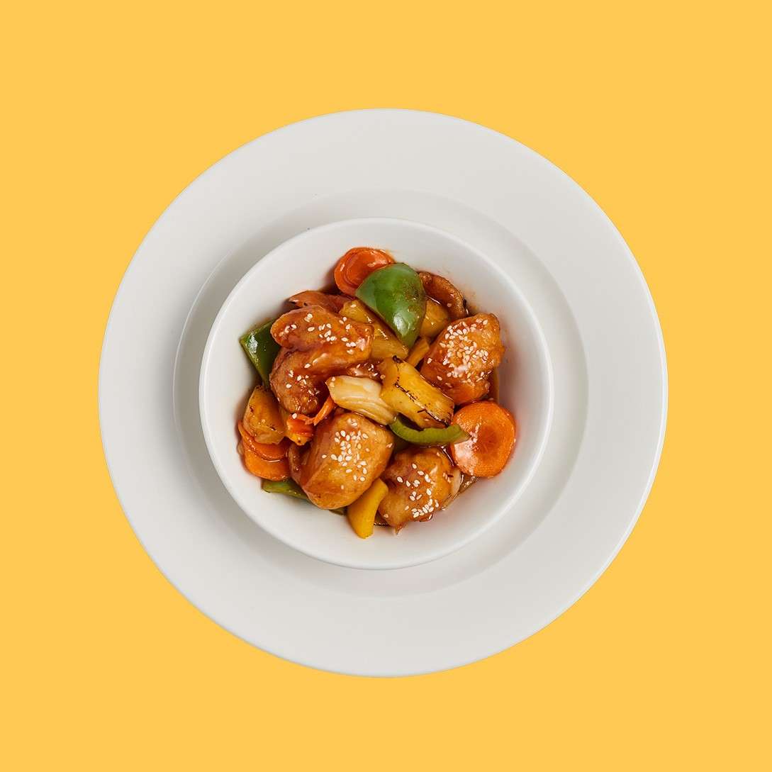 Bowl of glazed chicken pieces, topped with sesame seeds, is served with stir-fried bell peppers, carrots, and pineapple, set on white nested plates against a solid yellow background.