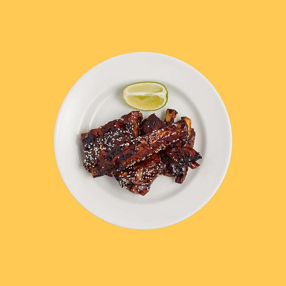 Glazed pork ribs rest on a white plate, sprinkled with sesame seeds, with charred edges and glossy sauce; a lime wedge sits beside them, set against a plain yellow background.
