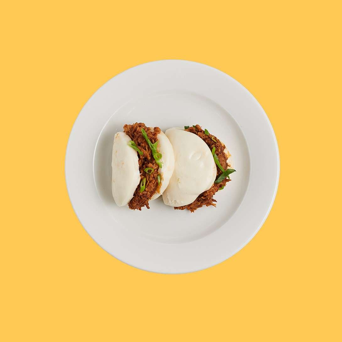Two steamed bao buns hold shredded meat and green onions, resting neatly on a white plate, centered against a plain yellow background.