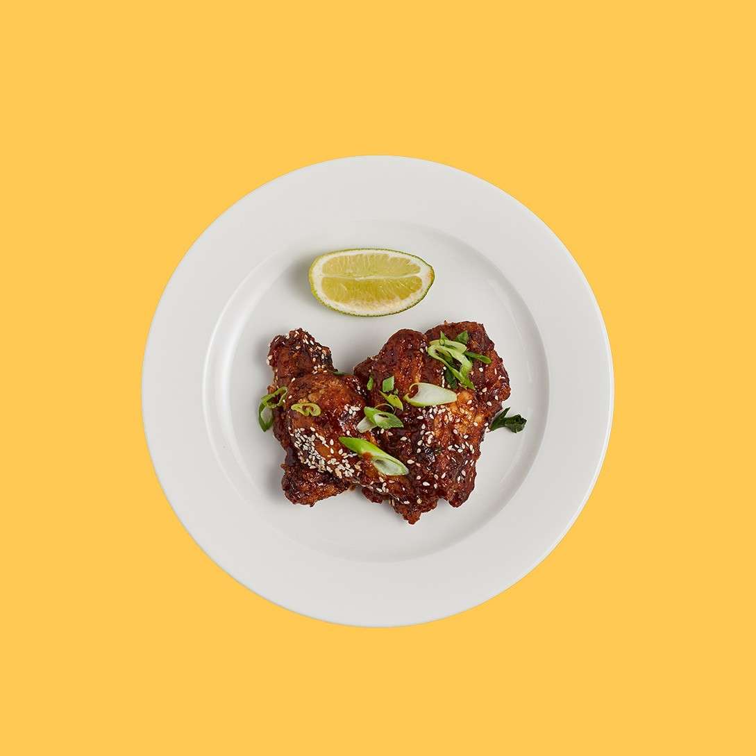 Chicken wings sit on a white plate, glazed and sprinkled with sesame seeds and sliced scallions, with a lemon wedge; plate isolated against a plain yellow background.