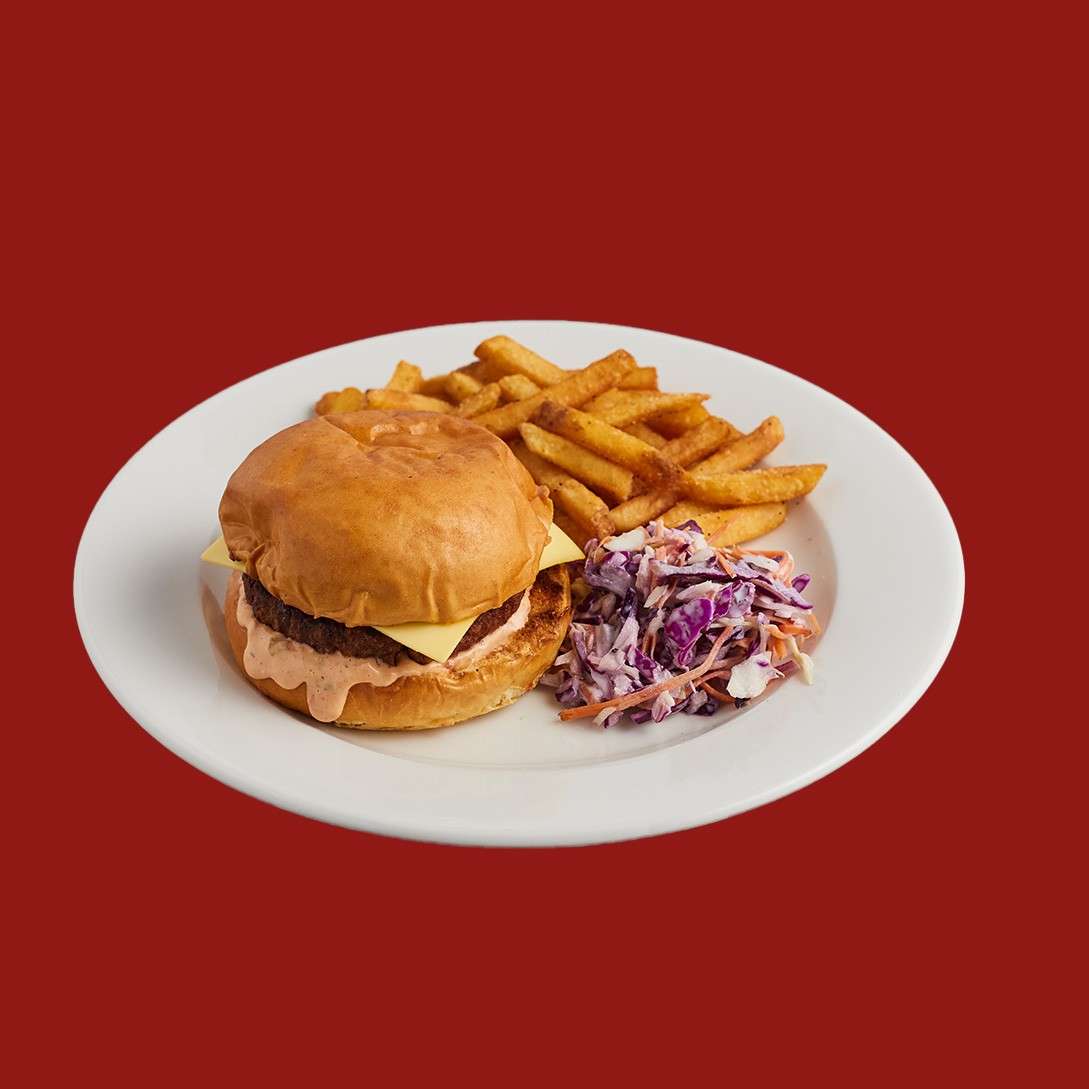 Chicken sandwich sits on a white plate, the grilled breast spilling from a soft bun; served with golden fries and purple-white coleslaw, set against a solid red background.