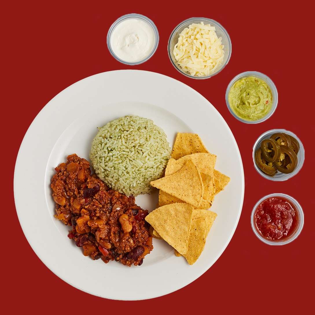 Mexican-style meal plated: green rice mound, bean chili, and tortilla chips on a white plate, surrounded by cups of sour cream, shredded cheese, guacamole, jalapeños, and salsa on red background.