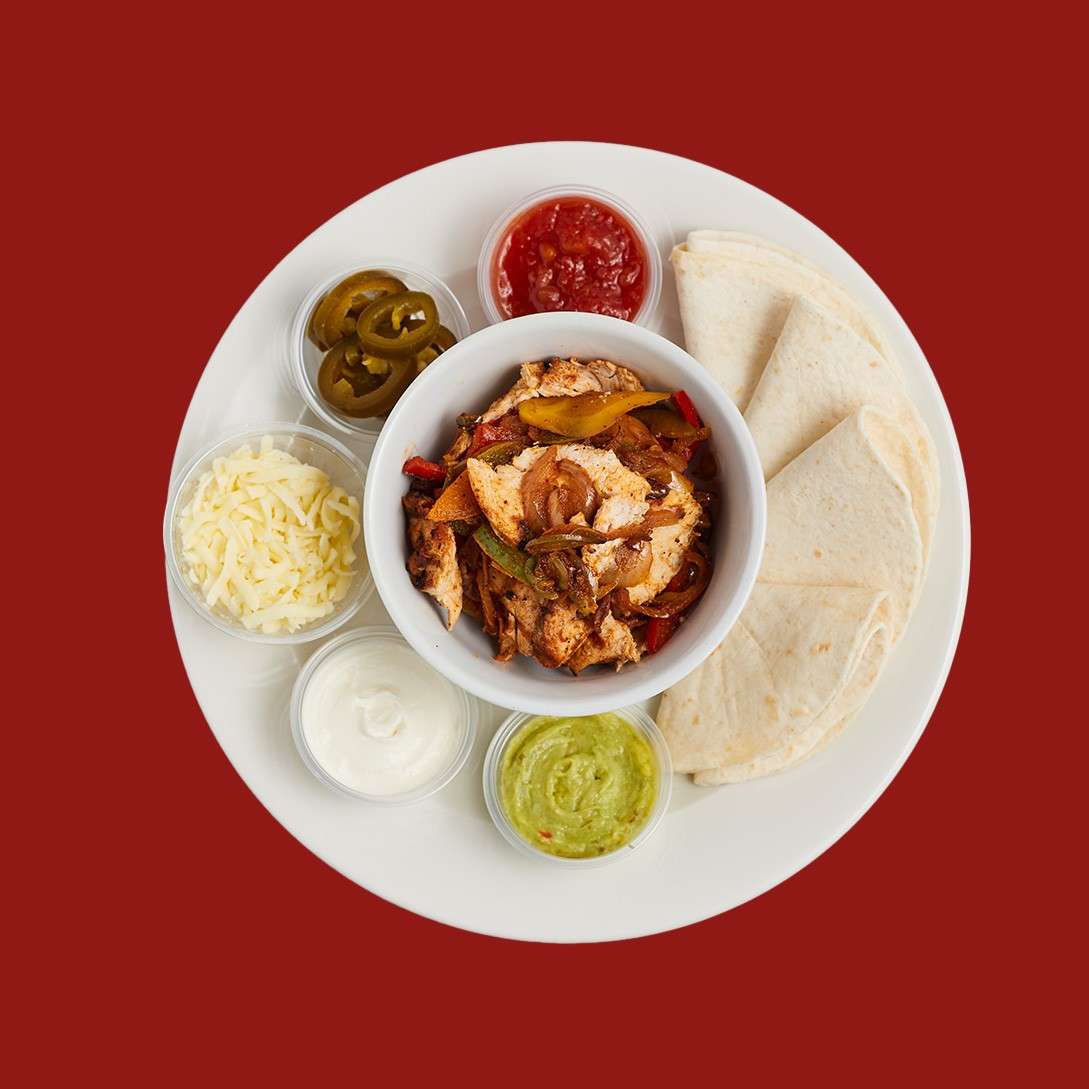 Chicken fajitas served; grilled chicken with peppers and onions fills a bowl, surrounded by tortillas, shredded cheese, sour cream, guacamole, salsa, and jalapeños on a white plate against red background.