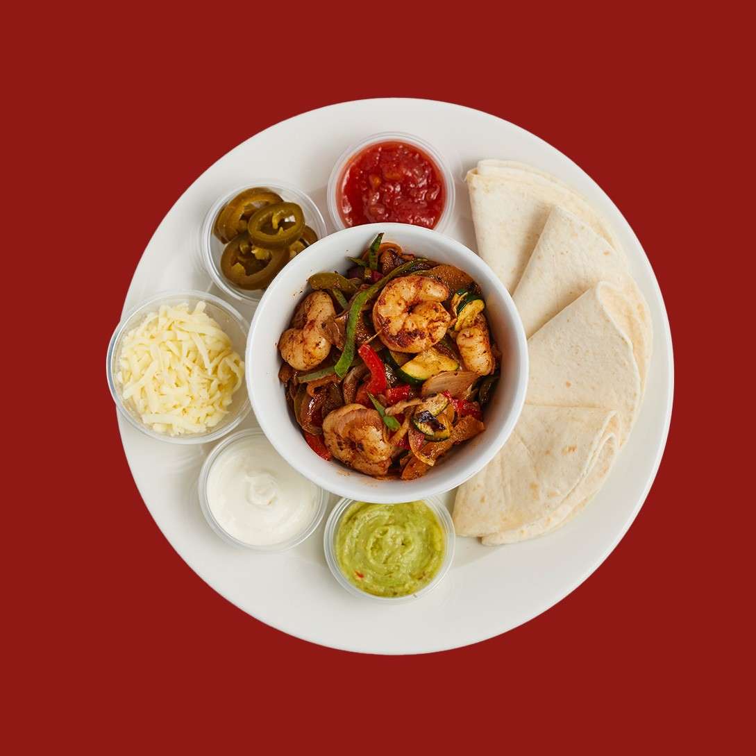 Bowl of seasoned chicken and vegetables sits centered. Around it, tortillas, shredded cheese, jalapeños, salsa, sour cream, and guacamole circle on a white plate against a red background.