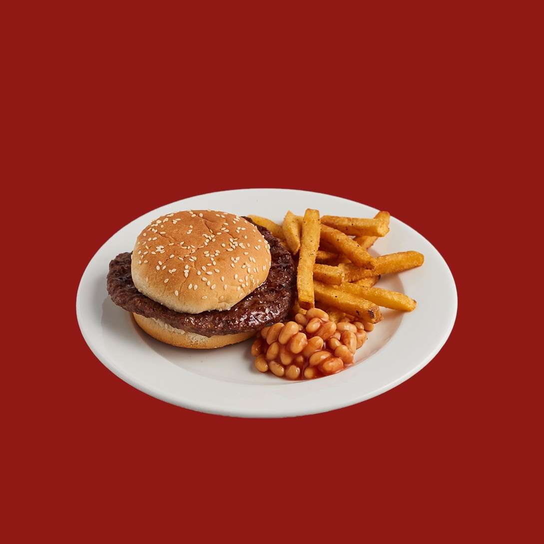 Burger rests on a white plate, accompanied by seasoned fries and baked beans; sesame bun holds a thin grilled patty. Context: centered against a solid red background.