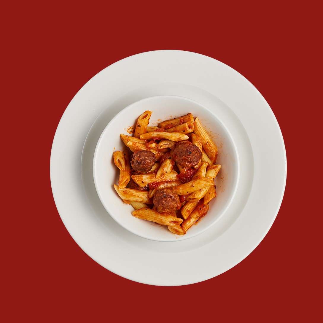 Penne pasta with meatballs in tomato sauce sits in a white bowl, centered on stacked white plates against a solid red background.
