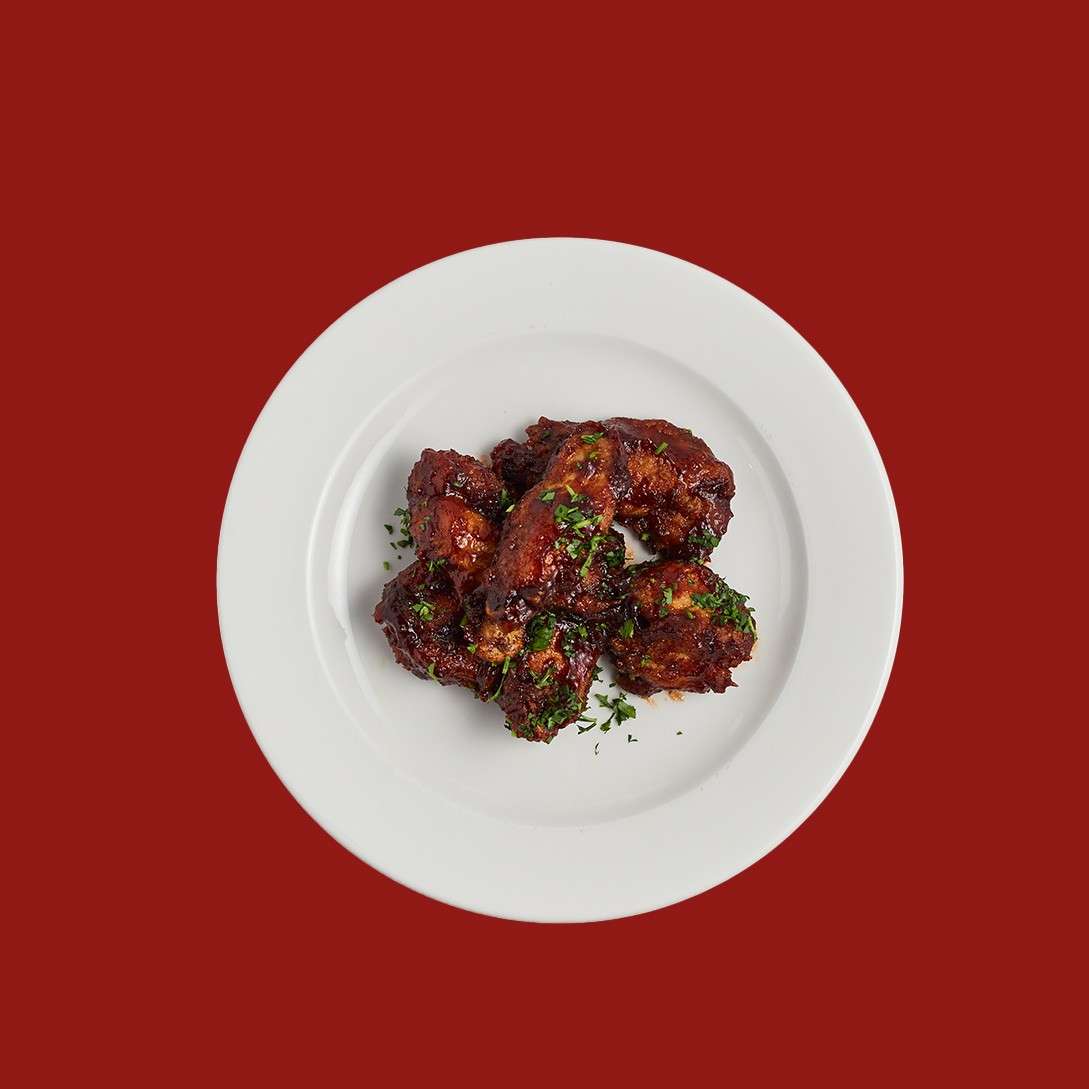 Glazed chicken wings rest in a small pile, sprinkled with chopped herbs, on a white plate set against a solid deep-red background.