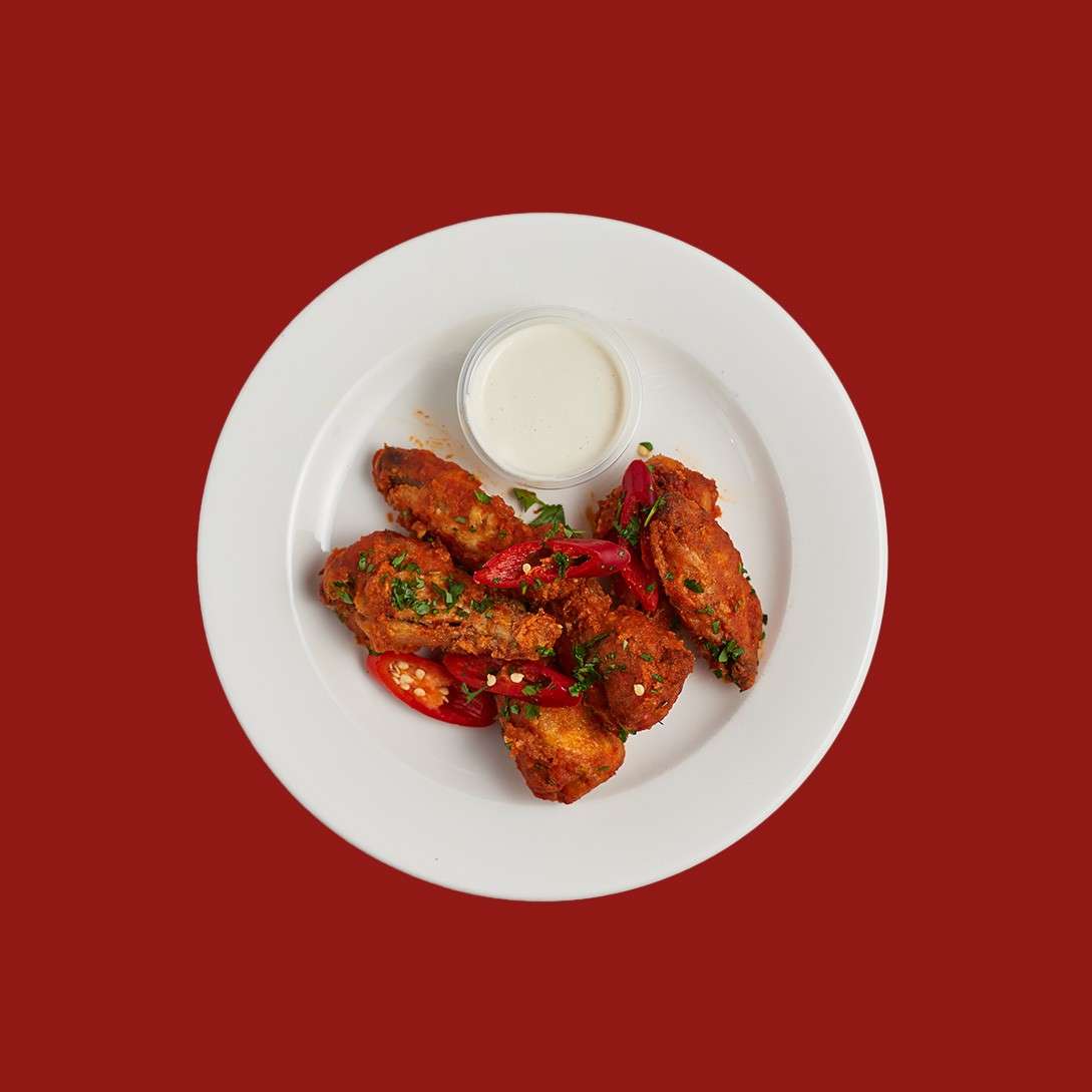 Red-seasoned chicken wings rest on a white plate, garnished with herbs and sliced chilies, beside a small cup of white dipping sauce, against a solid red background.
