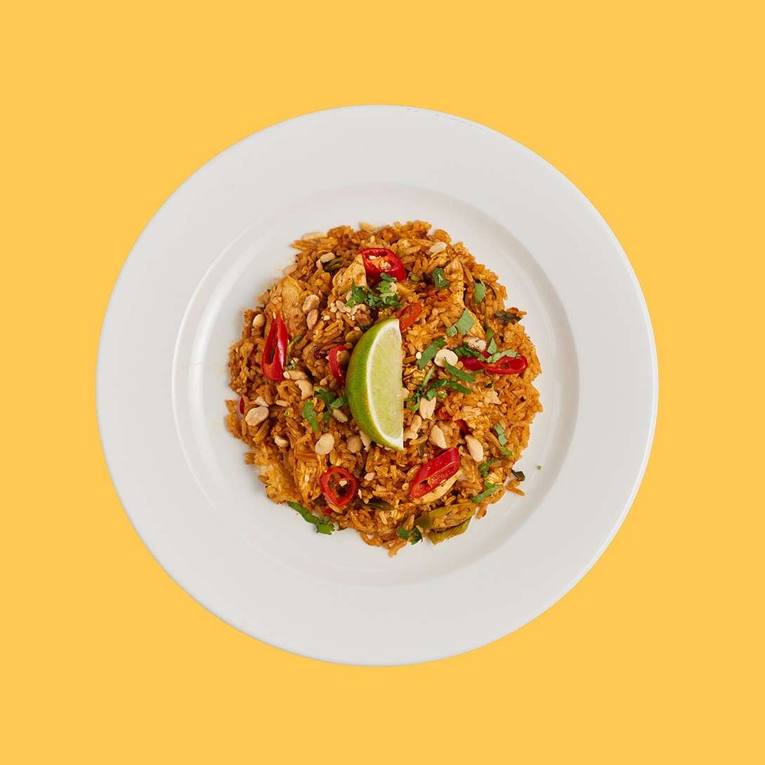 A mound of spicy fried rice sits garnished with red chili slices, peanuts, cilantro, and a lime wedge, served on a white plate against a yellow background.