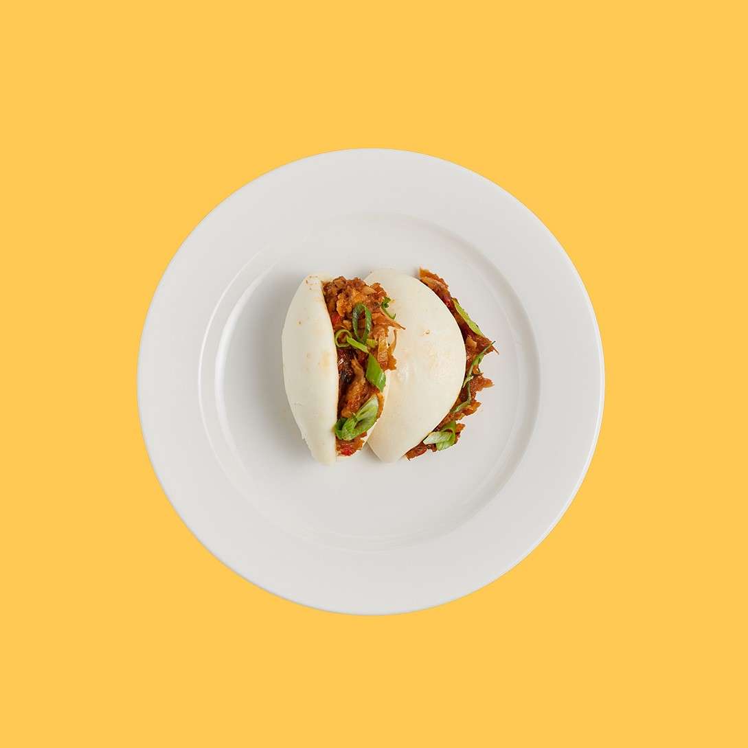 Two white steamed bao buns hold shredded meat and green onions, sitting centered on a white plate against a plain yellow background.