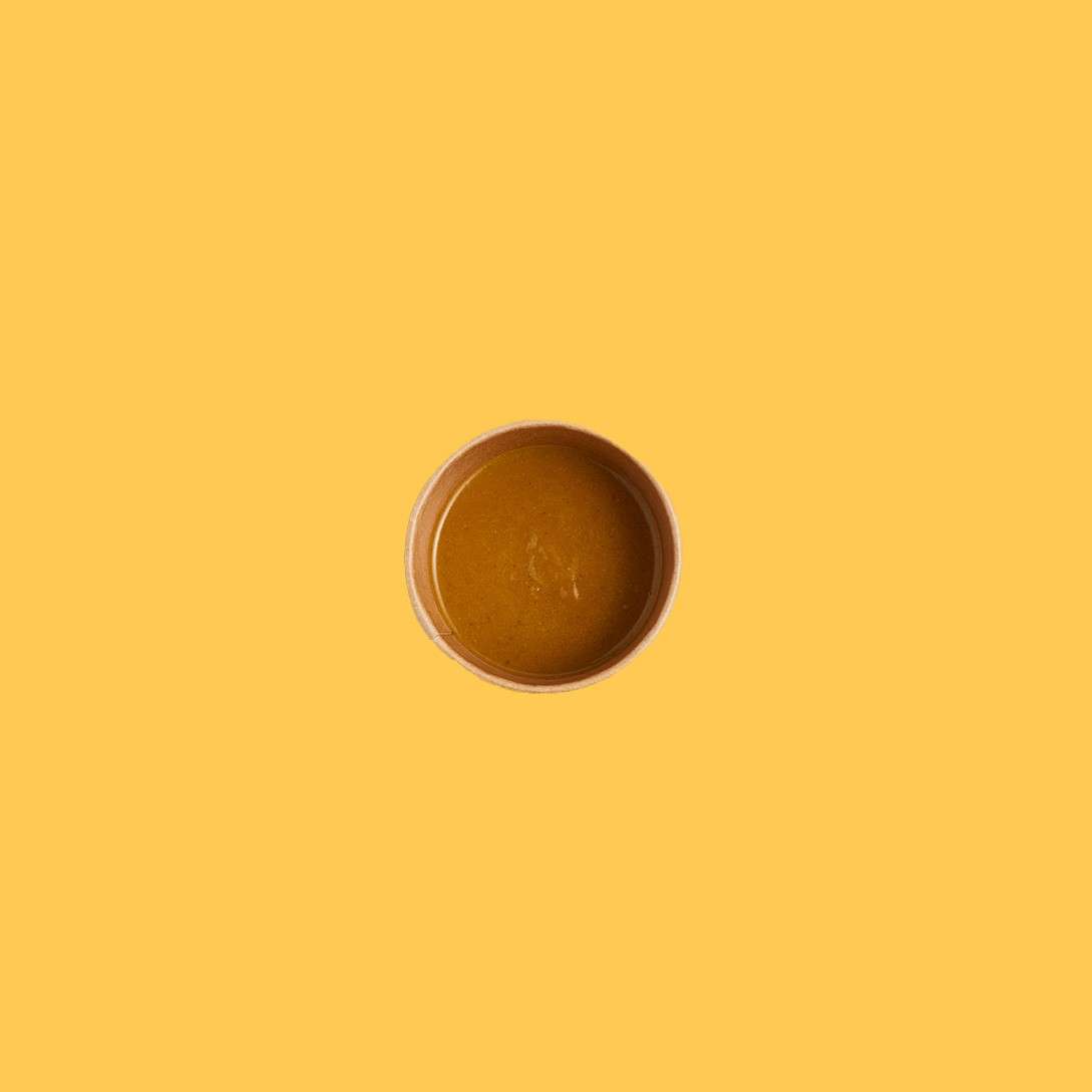 Small round bowl holds thick brown soup, viewed from above, resting alone against a flat mustard-yellow background, emphasizing minimalism and color contrast.