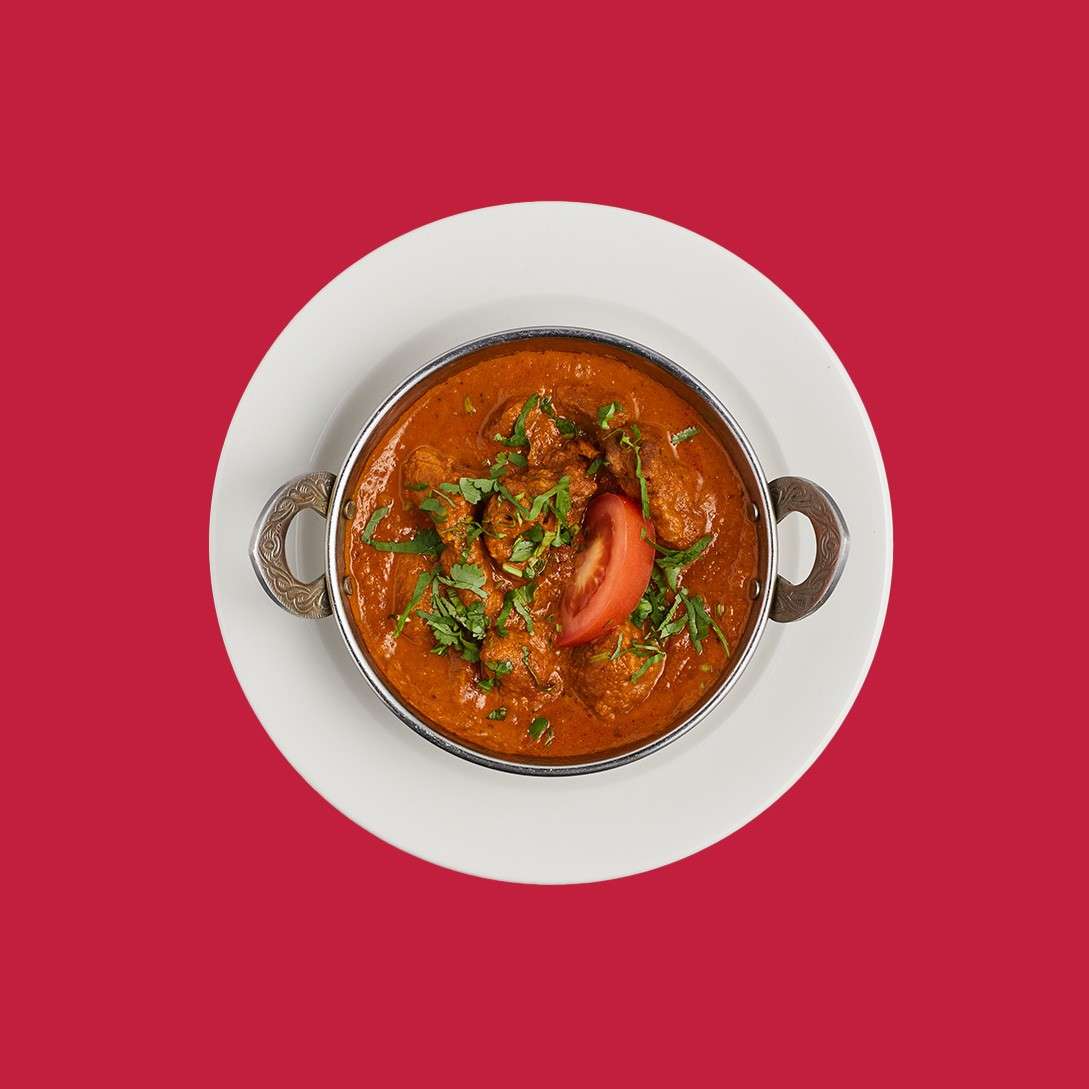 Metal-handled bowl of red curry with meat chunks, garnished with chopped cilantro and a tomato wedge, sits on a white plate, centered against a solid red background.