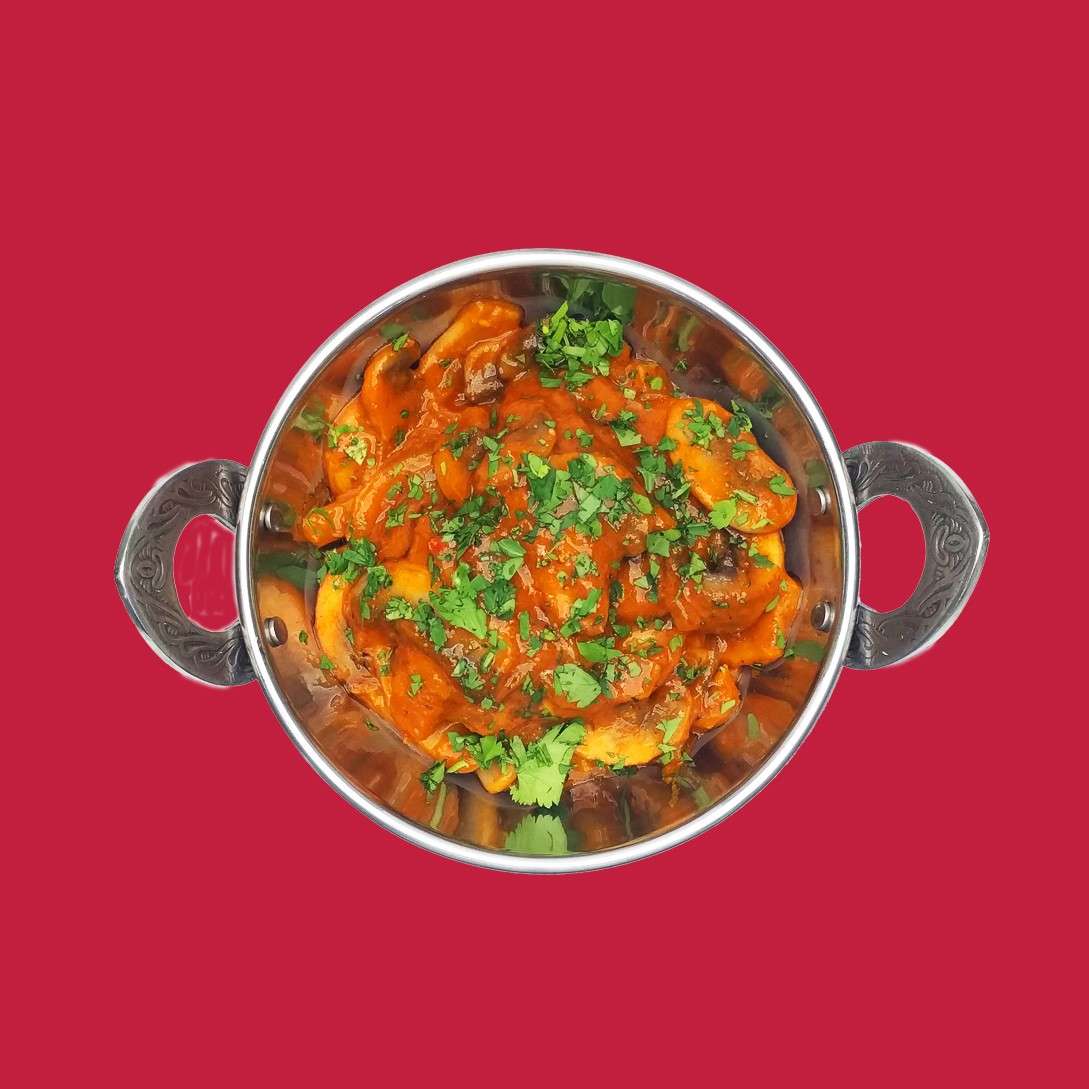 A metal serving bowl holds an orange-red saucy dish with visible vegetable pieces, garnished with chopped green herbs, centered against a plain solid red background.
