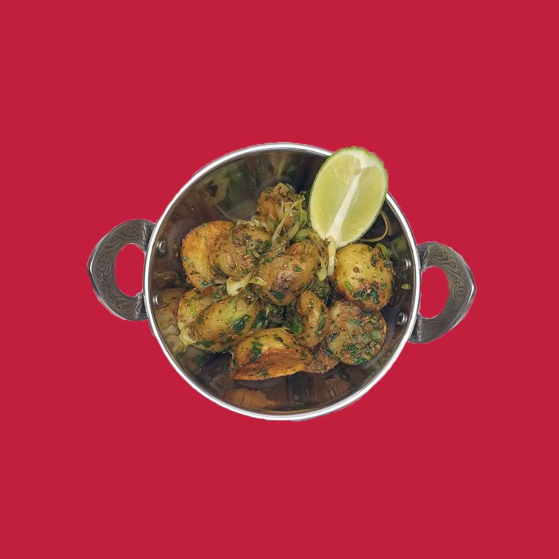 Spiced baby potatoes sit in a small metal handled bowl, tossed with chopped herbs and onion slices, topped with a lemon wedge, against a plain red background.