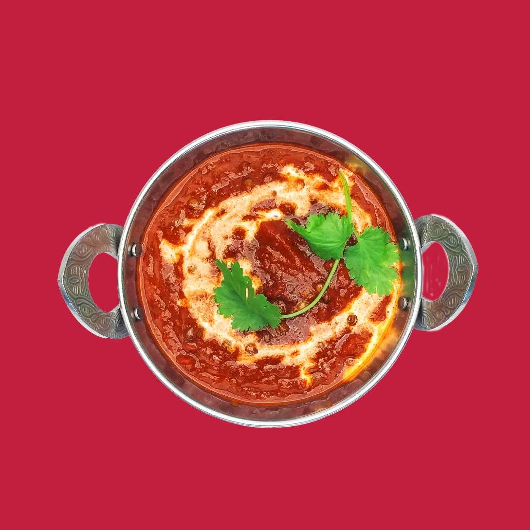 Two-handled metal bowl holds thick red curry, garnished with a cream spiral and cilantro leaves, resting centered against a solid red background, viewed from above.