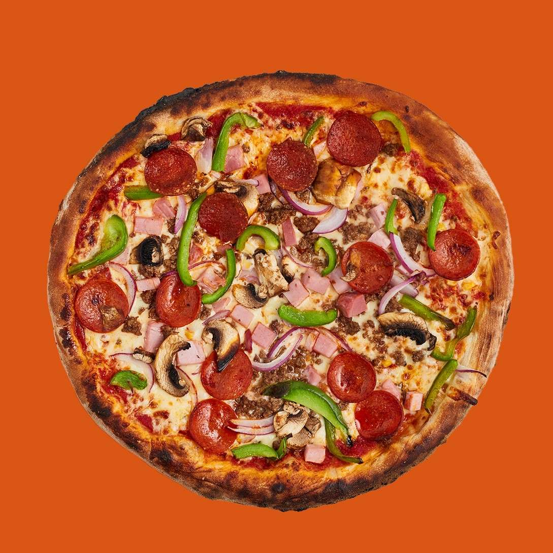 Whole pizza sits centered, topped with pepperoni, ham, sausage, mushrooms, green bell peppers, and red onion over cheese and tomato sauce, against a solid orange background.