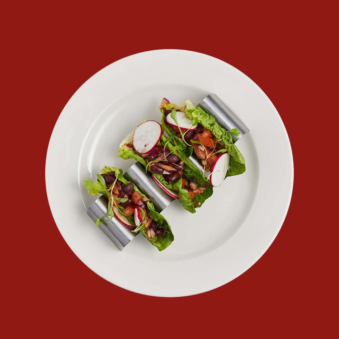 Three tacos rest in metal holders, filled with lettuce, black beans, radish slices, and salsa, arranged on a white plate against a solid red background.