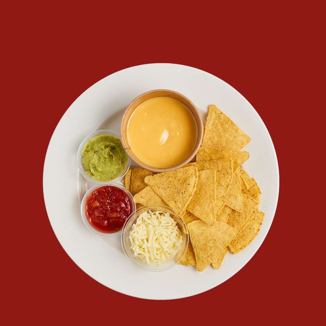 Tortilla chips sit beside dips—queso, guacamole, salsa, and shredded cheese—arranged for dipping on a white plate, set against a solid red background.