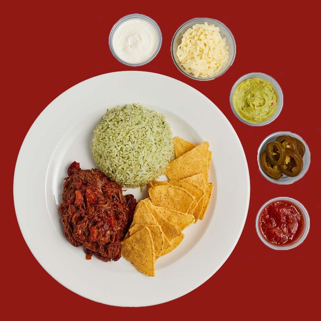Plate of Mexican-style meal: green herb rice, shredded beef in red sauce, and tortilla chips, with cups of sour cream, shredded cheese, guacamole, jalapeños, and salsa on a red background.