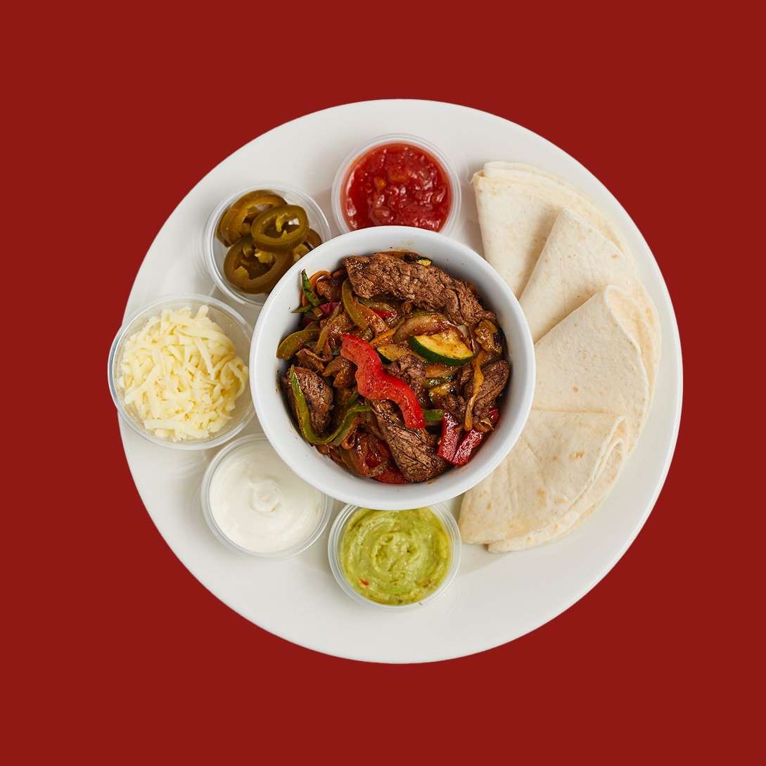 Bowl of sizzling beef fajitas with peppers sits centered, surrounded by tortillas and sides—salsa, jalapeños, shredded cheese, sour cream, guacamole—arranged on a white plate against a solid red background.