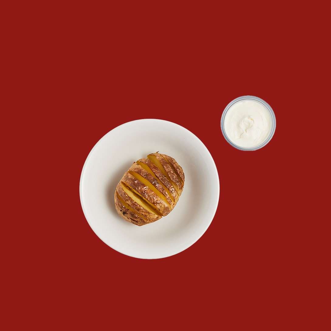 Bowl of mixed salad rests, featuring arugula, cherry tomatoes, red onion, and shredded carrots; a small cup of brown vinaigrette sits to the right, top-down on a solid red background.