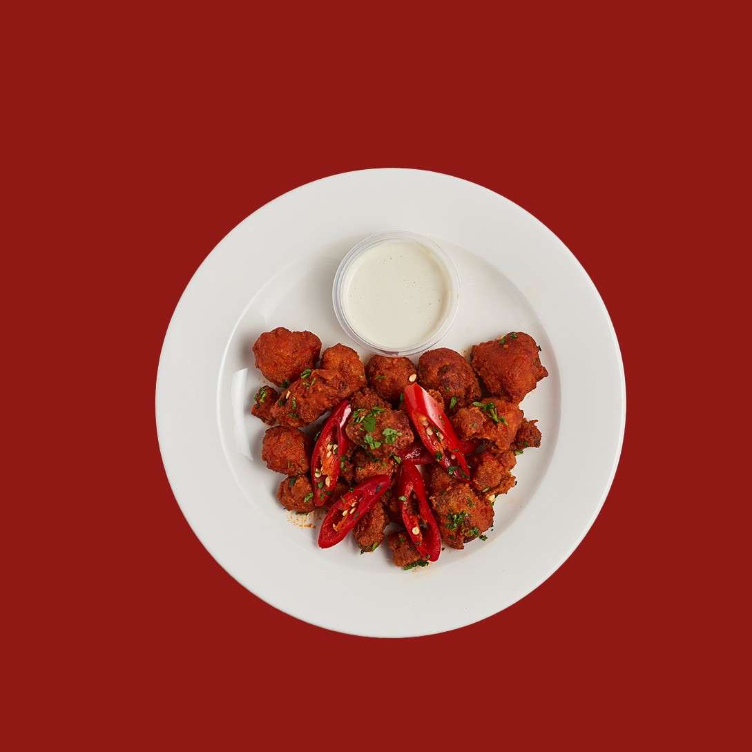 Fried bite-sized pieces sit with sliced red chiles, garnished with herbs, alongside a small cup of creamy dipping sauce, centered on a white plate against a solid red background.