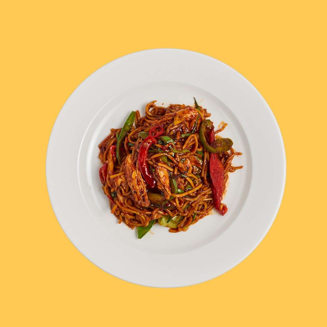 Stir-fried noodles tumble with sliced red and green peppers in glossy sauce, served on a white plate, centered against a solid yellow background.