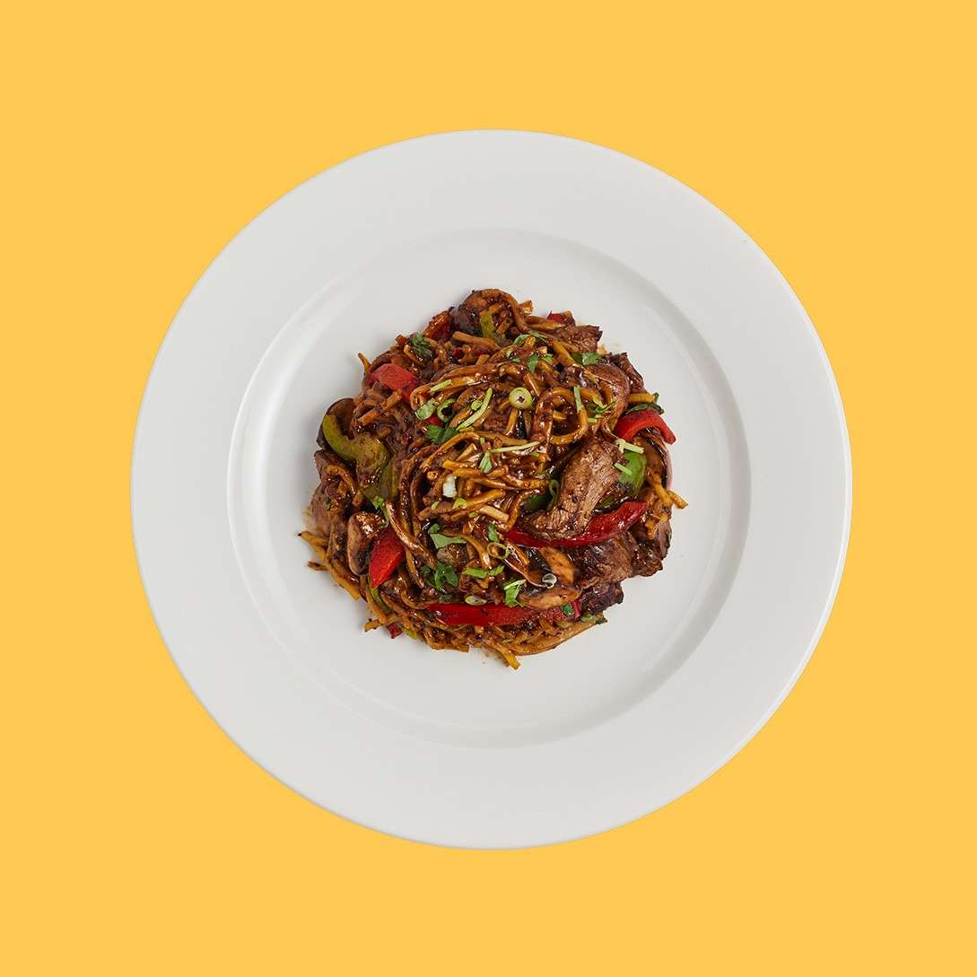 Stir-fried noodles with beef and vegetables sit heaped and glistening, garnished with scallions, on a white plate set against a solid yellow background.