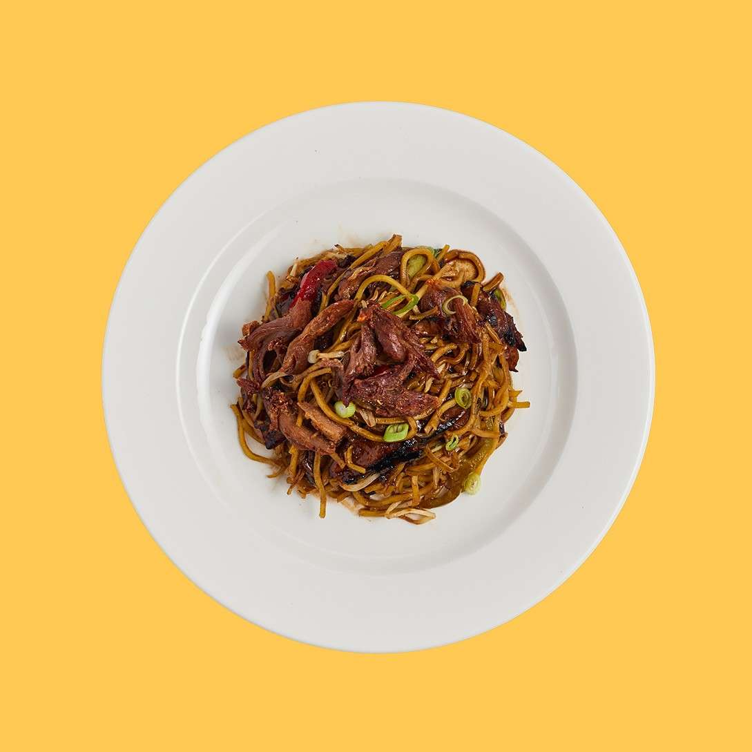Stir-fried noodles with sliced beef rest on a white plate, garnished with scallions, viewed overhead against a plain yellow background.