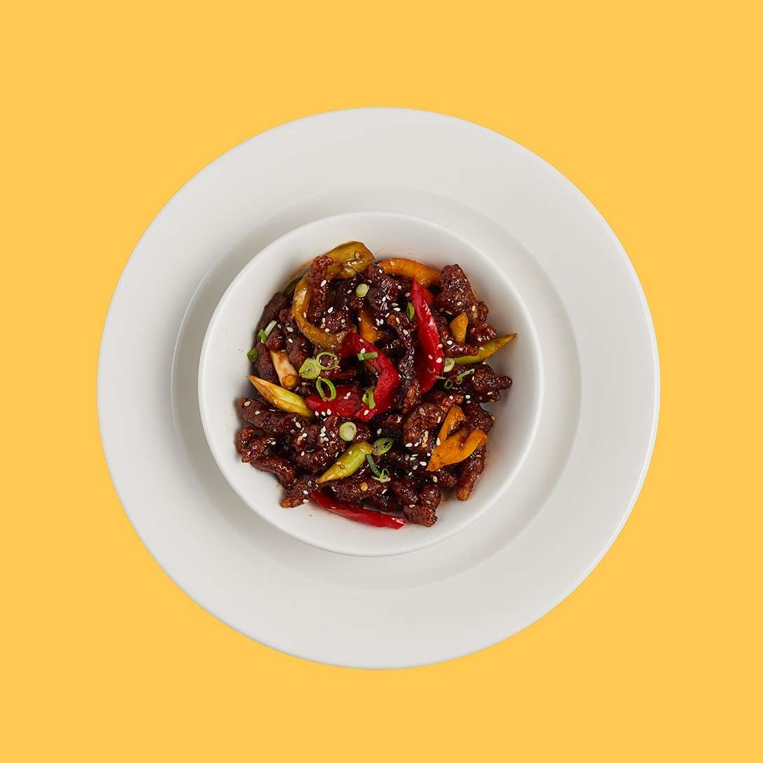 Bowl of glossy stir-fried beef and bell peppers, sprinkled with sesame seeds and scallions, sits centered on nested white plates against a flat yellow background.