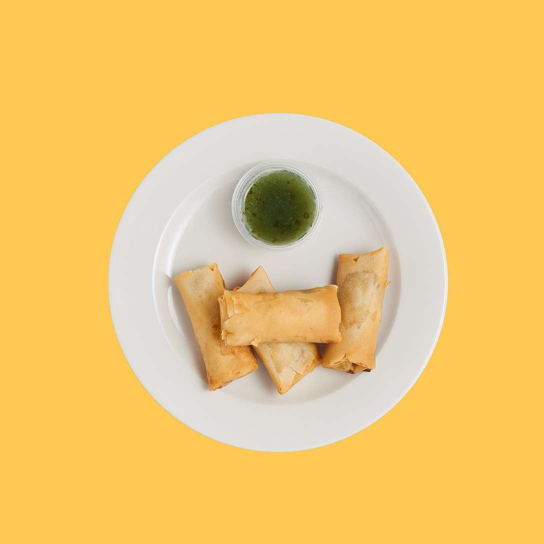Three golden spring rolls rest on a white plate with a small cup of green dipping sauce, centered against a solid yellow background.
