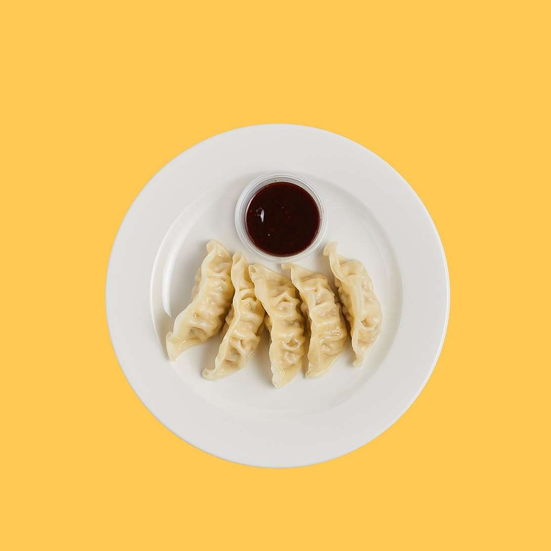 Five crescent dumplings encircle a cup of dark sauce on a white plate, set against a solid yellow background.