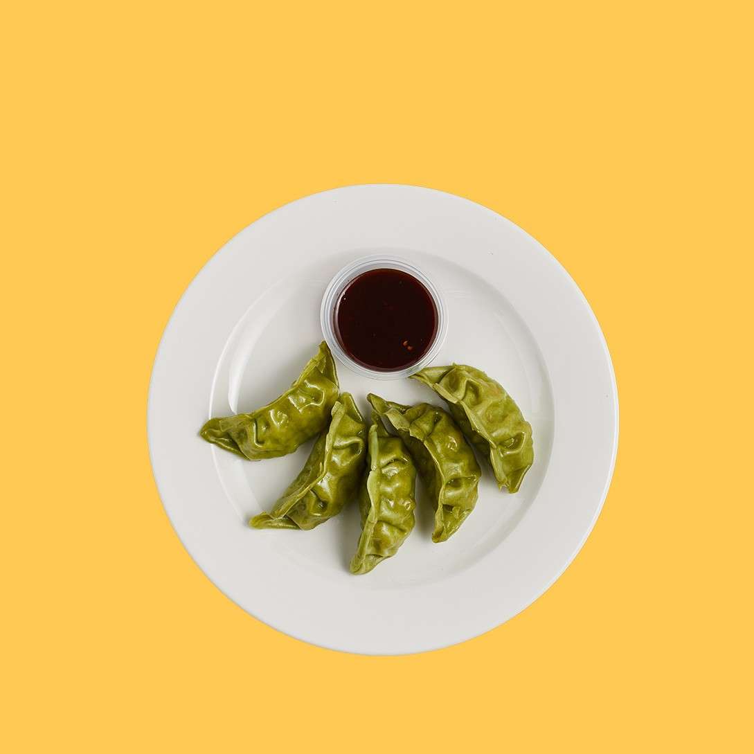 Green dumplings rest around a cup of dark sauce on a white plate, set against a solid yellow background.