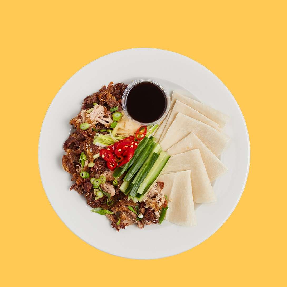 Plate of Peking duck components arranged neatly: shredded roast duck with crispy skin, green onion, red chili, cucumber sticks, and hoisin sauce; folded pancakes alongside, on a plate against a yellow background.