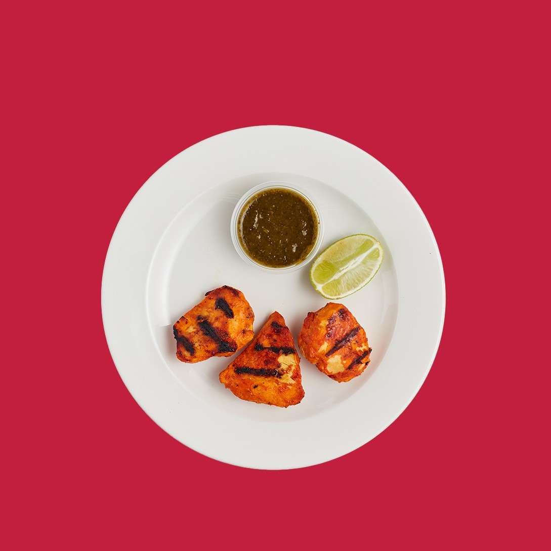 Three grilled marinated pieces rest on a white plate, accompanied by a small cup of green sauce and a halved lime; set against a solid red background.