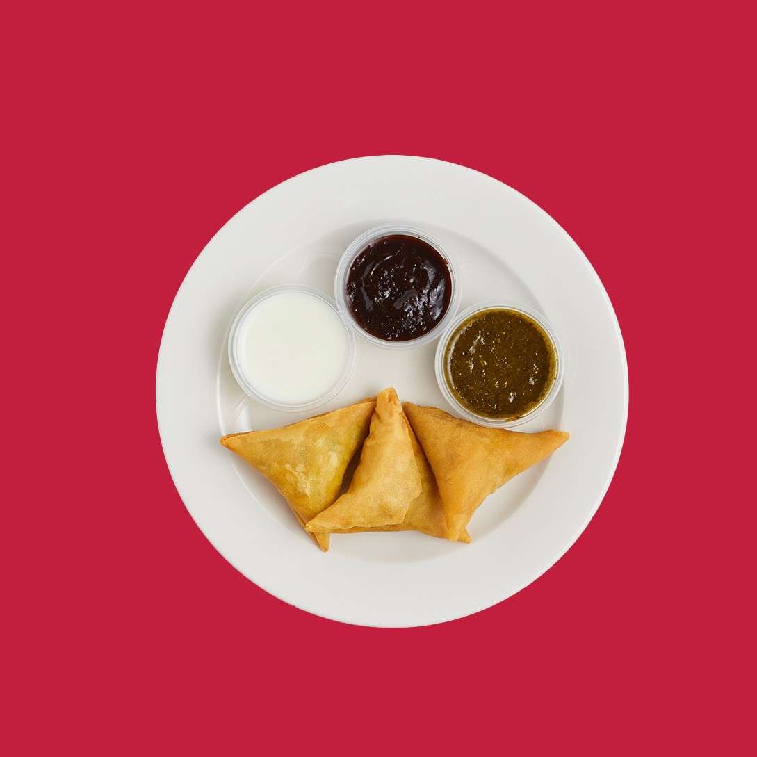 Three triangular fried pastries sit on a white plate, accompanied by three small dipping sauces—white, dark brown, and green—centered against a solid red background.