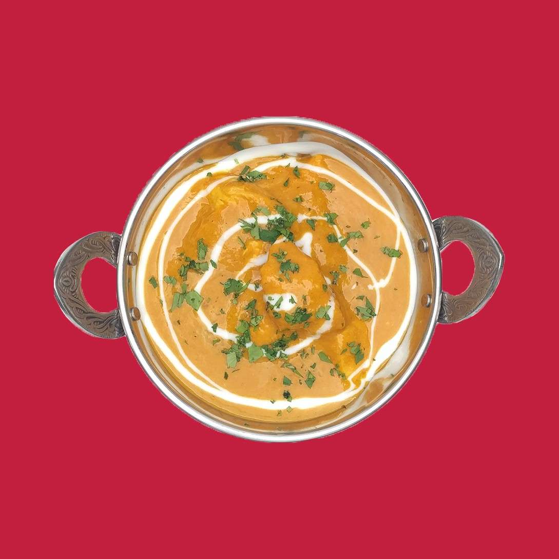Metal bowl of creamy orange curry, swirled with white cream and topped with chopped green herbs, sits centered. Handles flank the bowl. Context: top-down view against a plain red background.
