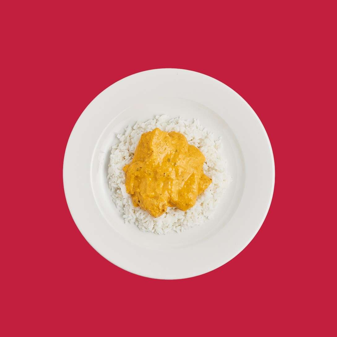 Plate of white rice topped with creamy orange curry rests centered; context: plain white plate on a solid red background, minimalist presentation with visible rice grains and smooth sauce.